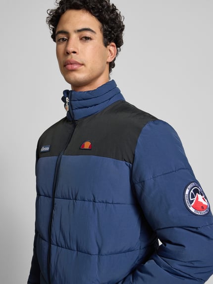 Männermodel trägt Steppjacke mit Reißverschluss Modell 'NEBULA' von Ellesse in Blau mit schwarzen Partien, Label-Details, Nahaufnahme Oberkörper. Regular Fit mit Stehkragen, Eingrifftaschen, dezente glänzende Textur, dezente Struktur auf Schulter und Brust.