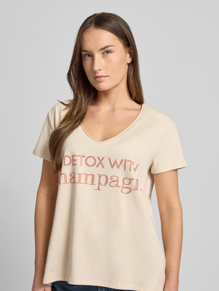 Frauenmodel trägt Loose Fit T-Shirt mit V-Ausschnitt von miss goodlife, beigefarbener Baumwoll-Elasthan-Mix, Statement-Print, Nahaufnahme Oberkörper.