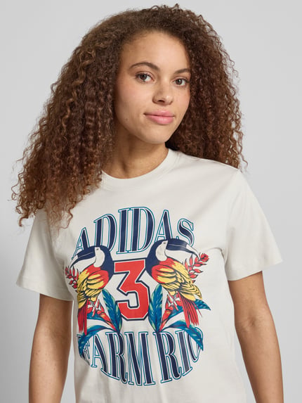 Frauenmodel trägt T-Shirt mit Label-Print von adidas Sportswear in Weiß, bunte Farben, Reine Baumwolle, Regular-Fit, Nahaufnahme Oberkörper.