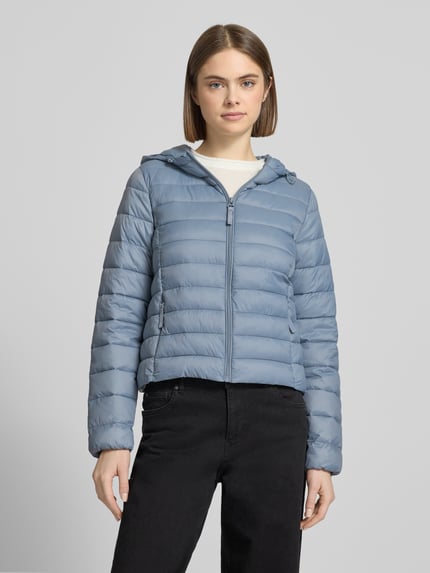 Modelka kobieca ma na sobie Regular Fit Steppjacke mit Kapuze Modell 'TAHIA' od Only, jasnoniebieska, jednobarwna, długi rękaw, stójka, elastyczny mankiet, boczne kieszenie, krój regular fit, zamek błyskawiczny, tekstura pikowana, widok całej sylwetki od przodu.