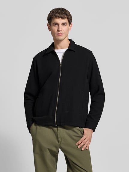 Männermodel trägt Wolljacke aus Woll-Mix von Forét in Schwarz, Umlegekragen, Reißverschluss, Regular Fit, Eingrifftaschen, Ganzkörperansicht von vorne.