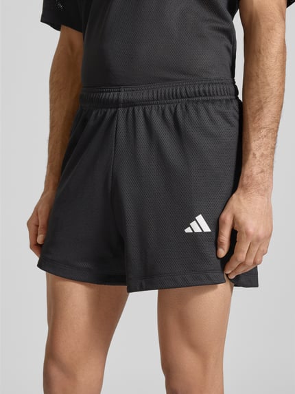 Mężczyzna nosi Straight Fit Shorts mit Logo-Print in Mesh-Optik od adidas Training, czarne krótkie spodenki, mesh, elastyczny pas, szerokość w rozmiarze M 39 cm, długość nogawki 12 cm, widok na nogi i talię, zbliżenie na dolną część ciała