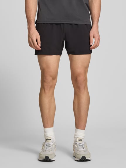 Mężczyzna model nosi Regular Fit Shorts mit Logo-Print od PUMA PERFORMANCE, czarny elastyczny materiał, krótkie spodenki, szerokość w rozmiarze M 38 cm, długość nogawki od wewnątrz 12 cm, widok całej sylwetki od przodu.
