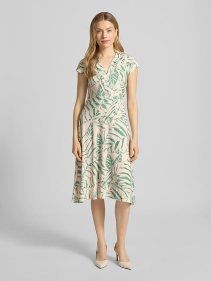 Modelka kobieca nosi Midikleid in Wickel-Optik mit V-Ausschnitt Modell 'Ca44rrara' od ZABAIONE, beżowo-zielony print liści, materiał elastyczny, krój slim fit, szerokość w rozmiarze S: 45 cm, długość tyłu: 105 cm, widok całej sylwetki z przodu.