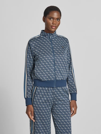 Frauenmodel trägt Regular Fit Sweatjacke mit Allover-Logo-Print von GUESS, blauer Ton mit weißem Muster, Reißverschluss, Nahaufnahme Oberkörper. Damen Sweatjacke, elastischer Material-Mix, Stehkragen, elastische Abschlüsse von GUESS Activewear.
