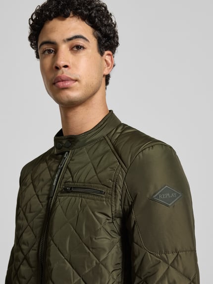 Männermodel trägt Regular Fit Jacke in Stepp-Optik von Replay in Olivgrün mit Stehkragen, Logo-Applikation, Nahaufnahme Oberkörper. Rückenlänge 66 cm, Schulterbreite 43 cm, Brustweite 52 cm, Ärmellänge 63 cm, Brusttasche und Eingrifftaschen mit Reißverschluss.