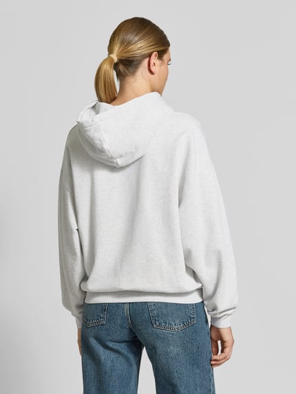 Frauenmodel trägt Oversized Hoodie aus reiner Baumwolle von Anine Bing in Hellgrau mit Känguru-Tasche, Label-Print und elastischen Abschlüssen, Ganzkörperansicht Rückansicht.