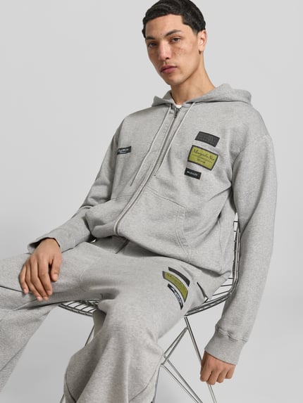 Męski model nosi Regular Fit Sweatjacke mit Logo-Patch od MM6 Maison Margiela z bawełny, krój regular fit, naszywka z logo, kaptur z tunelem, zamek błyskawiczny, długość tyłu w rozmiarze M: 69 cm, szerokość ramion w rozmiarze M: 45 cm, długość rękawów w rozmiarze M: 72 cm, jasnoszary kolor, tekstura gładka, elementy prostokątne, widok całego ciała z przodu.