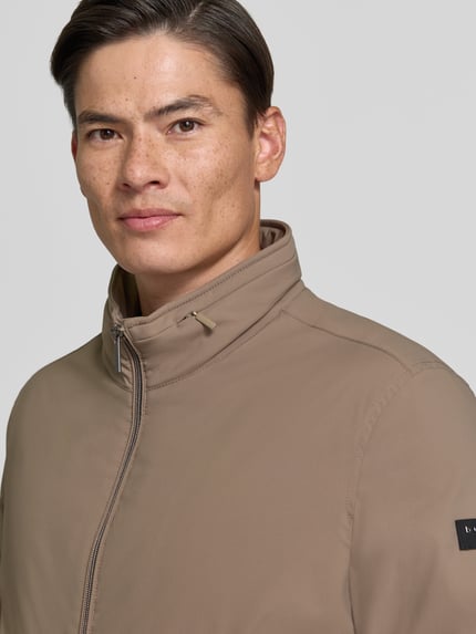 Männermodel trägt Regular Fit Jacke mit Stójka von bugatti w kolorze beżowym, zamek błyskawiczny, wpuszczane kieszenie, krój regular fit, elastyczny pas, długość tyłu w rozmiarze 50: 67 cm, szerokość ramion w rozmiarze 50: 45 cm, szerokość klatki piersiowej w rozmiarze 50: 58 cm, długość rękawów w rozmiarze 50: 67 cm, zbliżenie na górną część tułowia