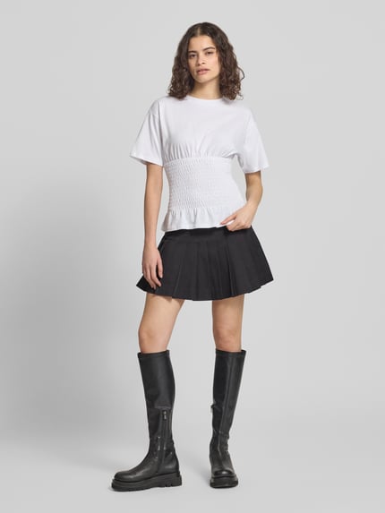 Vrouwenmodel draagt tailliertes T-Shirt met ronde hals, model 'EVELYN' van JJXX van zuiver katoen met smok-details, getailleerd fit, lengte achterzijde maat S: 53 cm, wit met zwarte plooirok, volledige lichaamsaanzicht van voren.
