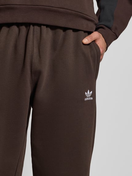 Mężczyzna nosi Straight Fit Sweatpants mit Logo-Stitching od adidas Originals, brązowa bawełna, elastyczny pas, detal logo, zbliżenie na biodra.