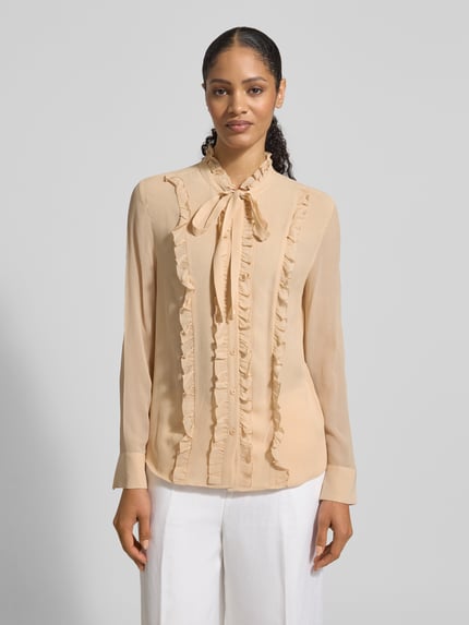 Vrouwenmodel draagt Regular Fit Bluse aus Seide mit Rüschen van HERZENS in lichtbeige met ruches, knoopsluiting, strik, pure zijde, regular fit, ruglengte 64 cm, mouwlengte 63 cm, voorkant hele lichaam zichtbaar.