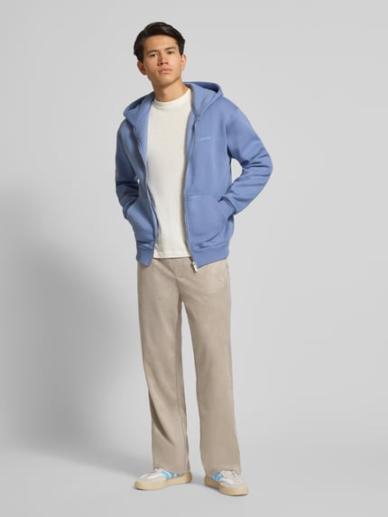 Männermodel draagt Stoffhose mit elastischem Bund van Pegador in beige met los vallende pasvorm, Franse steekzakken, elastische tailleband, normale lengte, Ganzkörperansicht von vorne.