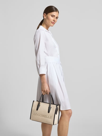 Frauenmodel trägt helle Handtasche mit Label-Stitching von Coccinelle, Hauptfach mit Reißverschluss, Tragehenkel, Schulterriemen, Breite 27 cm, Länge 20 cm, Tiefe 13 cm, Länge Schulterriemen 105 cm; Ganzkörperansicht von vorne.