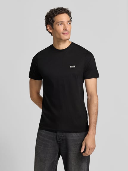 Męski model nosi Slim Fit T-Shirt mit Rundhalsausschnitt od GUESS, czysta bawełna, krój slim fit, naszywka z logo, czarny kolor, długość tyłu w rozmiarze M: 70 cm, widok całej sylwetki z przodu