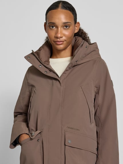 Frauenmodel trägt Regular Fit Parka mit Kapuze Modell 'Melody' von Didriksons in Braun, glatte Struktur, aufgesetzte Taschen, Nahaufnahme Oberkörper. Rückenlänge 111 cm, Schulterbreite 42 cm, Brustweite 53 cm, Ärmellänge 65 cm.