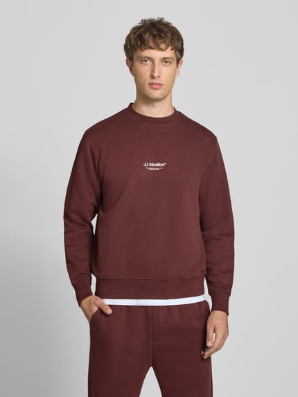 Männermodel trägt Regular Fit Sweatshirt aus Baumwoll-Mix Modell 'Soho' von JACK & JONES in Bordeaux mit Logo-Print, Ganzkörperansicht von vorne.