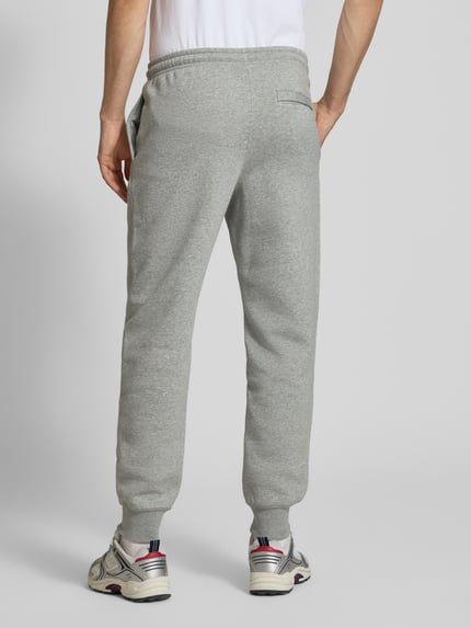 Mężczyzna nosi spodnie dresowe Sweatpants mit Label-Stitching und Gesäßtasche von Nike, mieszanka bawełny, krój regular fit, wyhaftowane logo, elastyczny pas, kieszeń z tyłu, długość wewnętrzna nogawki w rozmiarze M: 76 cm, jasnoszary melanż, miękka faktura, gładkie powierzchnie, całkowity widok od tyłu.