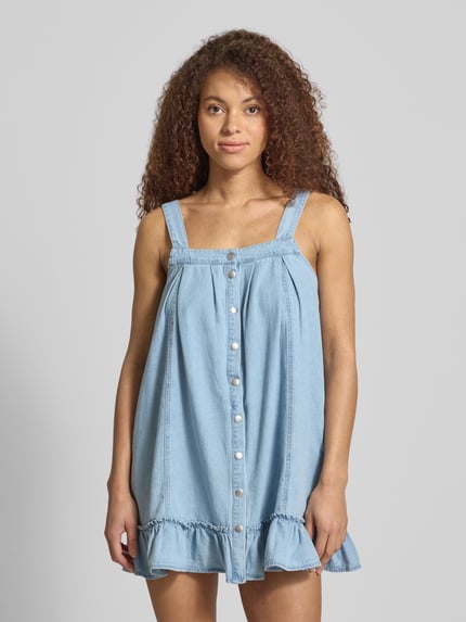 Frauenmodel trägt Jeanskleid aus reiner Baumwolle Modell 'OREGON' von Noisy May in Hellblau mit Rüschen und Druckknöpfen, Ganzkörperansicht von vorne.