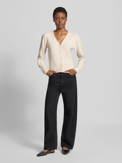 Frauenmodel trägt Regular Fit Strickjacke mit V-Ausschnitt Modell 'DOFFY' von VERO MODA in Beige und schwarze Stoffhose, Ganzkörperansicht von vorne. Damen Strickjacke von VERO MODA.