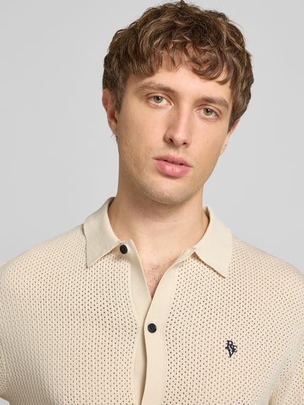 Męski model nosi Slim Fit Strick-Poloshirt mit Label-Stitching od REVIEW z mieszanki wiskozy, paski w kontrastowym kolorze, listwa guzikowa, wyhaftowane logo, długość tyłu w rozmiarze M: 65 cm, jasnobeżowy kolor, dzianinowa faktura, widoczny wzór, Nahaufnahme Oberkörper.