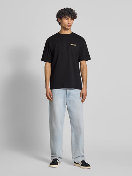 Männermodel trägt ein unifarbenes, schwarzes Relaxed Fit T-Shirt aus reiner Baumwolle Modell 'FRED DU MARTIN' von Only & Sons mit Motiv-Print, Rundhals und kurzen Ärmeln, feine Rippenstruktur, Ganzkörperansicht von vorne.