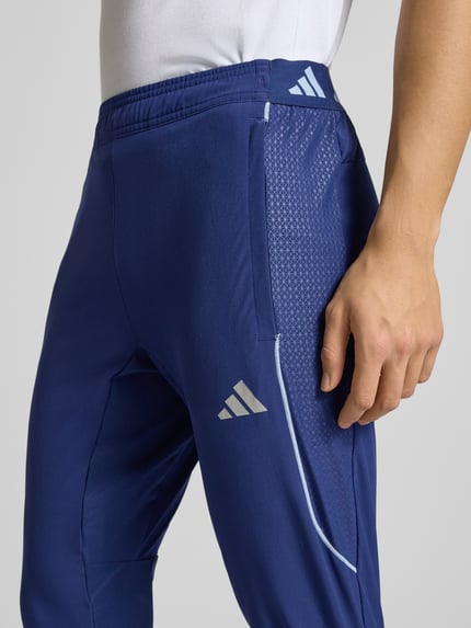 Männermodel draagt Slim Fit Sweatpants met Logoprint model 'Tech' van adidas Training in blauw met elastische materiaalmix, detailopname.