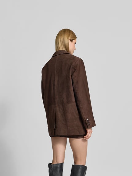 Modelka kobieca nosi Blazer aus echtem Ziegenvelours od ROTATE, brązowy z kołnierzem z klapami, oversize, listwa guzikowa, kieszenie z patkami, długość pleców w rozmiarze 36: 77 cm, szerokość klatki piersiowej w rozmiarze 36: 60 cm, długość rękawa w rozmiarze 36: 64 cm, szerokość talii w rozmiarze 36: 58 cm, miękka faktura zamszowa, widok całej sylwetki od tyłu.