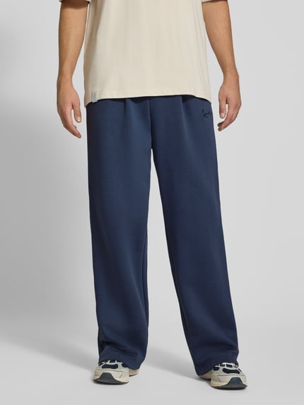 Männermodel trägt Sweatpants mit Logo-Stitching und Eingrifftaschen von Karl Kani in Dunkelblau, Baumwoll-Mix, Wide Fit, Ganzkörperansicht von vorne.