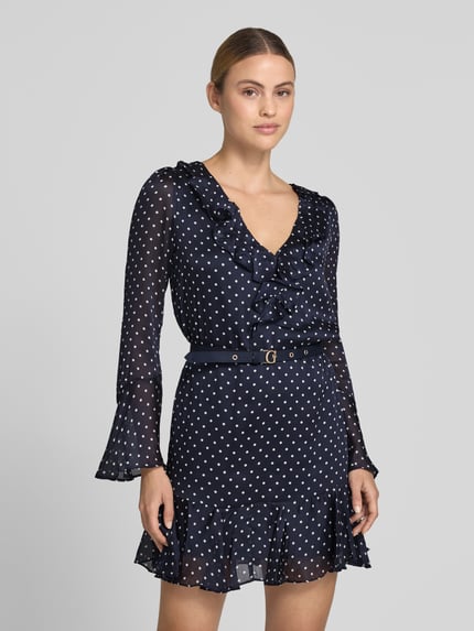Frauenmodel trägt Minikleid mit Rüschenbesatz von GUESS in Dunkelblau mit weißen Punkten, Taillierte Passform, Abnehmbarer Taillengürtel, V-Ausschnitt, Langärmelig, Rückenlänge bei Größe S 83 cm, Brustweite 42 cm, Ärmellänge 62 cm, Ganzkörperansicht von vorne.
