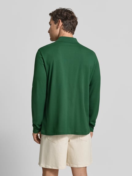 Männermodel trägt Regular Fit Poloshirt aus reiner Baumwolle von Lacoste in Dunkelgrün, helle Shorts, Ganzkörperansicht Rückansicht.