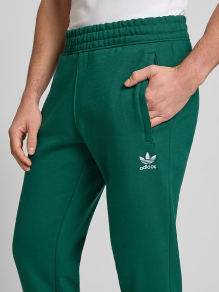 Mężczyzna nosi Sweatpants mit elastischem Bund od adidas Originals, zielony bawełniany materiał, luźny krój, kieszenie, haft, zbliżenie na biodra