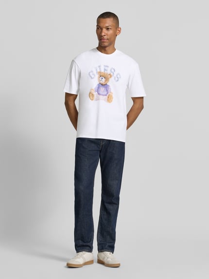 Männermodel trägt T-Shirt mit Motiv-Print und Rundhalsausschnitt von Guess, Herren T-Shirt aus reiner Baumwolle, Regular-Fit, Hauptfarbe Weiß, Motiv mit Bär und Logo, Rückenlänge bei Größe M 68 cm, Ganzkörperansicht von vorne.