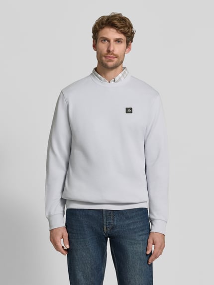 Męski model trägt Sweatshirt mit Label-Patch von Scotch & Soda aus bawełna, luźny krój, okrągły dekolt, prążkowane wykończenia, długość tyłu w rozmiarze M 64 cm, długość rękawa M 64 cm, jasnoszary, kształt prosty, faktura gładka, Ganzkörperansicht von vorne.