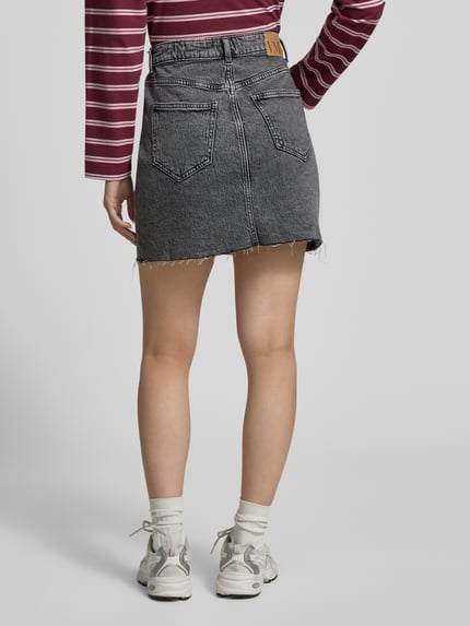 Frauenmodel trägt Regular Fit Jeansrock aus Baumwoll-Mix Modell 'TESSA' von Vero Moda in Denim-Optik, Mini-Länge, Label-Patch, 5-Pocket-Design, Materialverarbeitung Twill, Ganzkörperansicht Rückansicht.