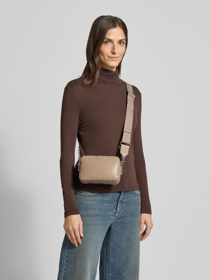 Frauenmodel trägt Crossbody Bag mit Logo-Prägung Modell 'Webbing' von CK Calvin Klein in Leder-Optik, Hauptfach mit Reißverschluss, verstellbarer Tragehenkel, Farbe Taupe, Breite 18 cm, Länge 12 cm, Tiefe 6 cm, Länge Schulterriemen 75 cm, Ganzkörperansicht von vorne.