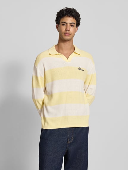 Męskimodel nosi Regular Fit Strick-Poloshirt mit Block-Streifen von REVIEW z bawełny, krój regular fit, blokowe pasy, haftowane logo na piersi, długość tyłu w rozmiarze M 66 cm, długość rękawów 61 cm, jasne żółte i białe pasy z prążkowaną fakturą, widok całego ciała z przodu.