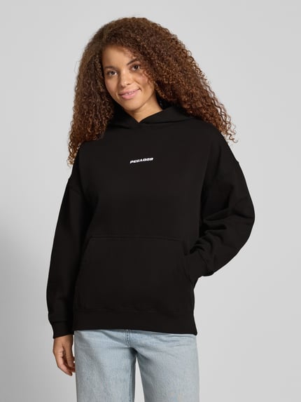 Frauenmodel trägt Oversized Hoodie mit Label-Stitching Modell 'CLARITA' von Pegador in Schwarz, Baumwoll-Mix, Nahaufnahme Oberkörper.