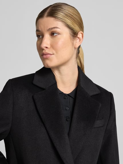 Frauenmodel trägt Regular Fit Mantel mit Reverskragen Modell 'DALI' von MANGO in Schwarz, glatte Textur, Nahaufnahme Oberkörper. Rückenlänge 93 cm, Schulterbreite 38 cm, Brustweite 47 cm, Ärmellänge 60 cm.