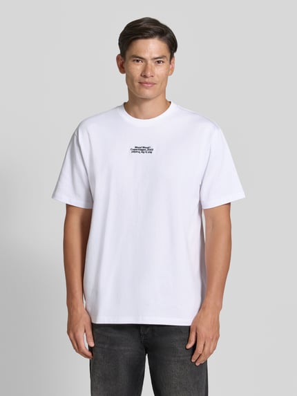 Männermodel trägt Regular Fit T-Shirt aus reiner Baumwolle Modell 'Oliver' von Wood Wood, schlichtes Weiß, Logo-Stitching, Ganzkörperansicht von vorne. Rückenlänge bei Größe M: 73 cm.
