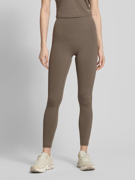Frauenmodel trägt Leggings mit elastischem Bund und Logo von P.E Nation, braun, Material-Mix mit Stretch-Anteil, High-Rise, körpernahe Passform, Ganzkörperansicht von vorne.