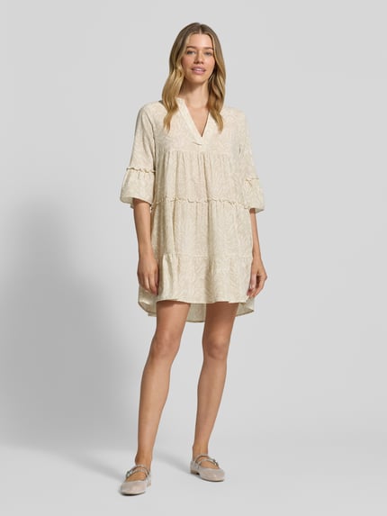 Modelka kobieca nosi Ausgestelltes Blusenkleid aus reiner Baumwolle Modell 'DICTHE' od Vero Moda, kremowa mini sukienka z bawełny z falbanami, wzór allover, rękaw 1/2 trąbkowy, widok całej sylwetki z przodu.