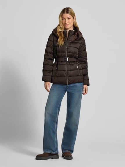 Modelka kobieca nosi Steppjacke mit Reißverschlusstaschen und Gürtel von monari, brązowa, krój slim fit, kaptur z ekofutrem, dwukierunkowy zamek, pasek, długość tyłu w rozmiarze 36: 67 cm, szerokość ramion w rozmiarze 36: 42 cm, szerokość w biuście w rozmiarze 36: 49 cm, długość rękawa w rozmiarze 36: 62 cm, pełny widok z przodu.