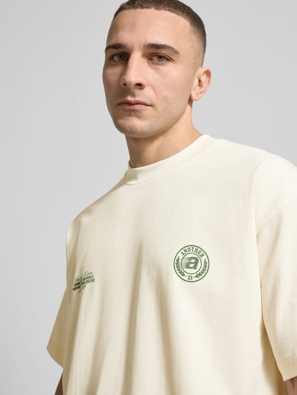 Männermodel trägt T-Shirt aus Baumwolle Modell 'Sunday Miles' von ANOTHER COTTON LAB in Creme mit grünen Label-Details, Nahaufnahme Oberkörper.