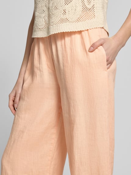 Frauenmodel trägt Leinenhose mit elastischem Bund und Eingrifftaschen von Emily Van den Bergh in Pfirsich, Loose Fit, Nahaufnahme Oberkörper, Reines Leinen, französische Eingrifftaschen, angedeutete Gesäßleistentaschen, Struktur leicht texturiert.
