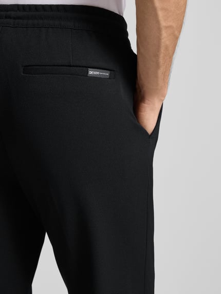 Mężczyzna nosi czarne spodnie Tapered Chino mit Viskose-Anteil od Tom Tailor Denim; detal tylnej kieszeni, tekstura gładka, zbliżenie tyłu.