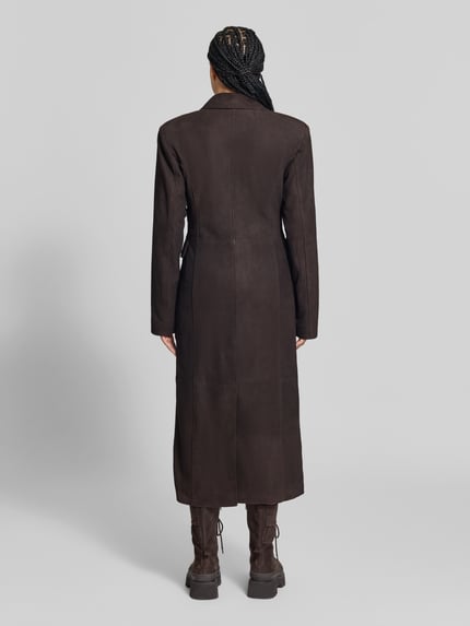 Frauenmodel trägt Ledermantel aus echtem Leder von ROTATE in Dunkelbraun, strukturierte Velours-Oberfläche, tailliert, Ganzkörperansicht Rückansicht. Rückenlänge 129 cm, Brustweite 42 cm, Ärmellänge 66 cm, Doppelreiher mit Reverskragen und Klappentaschen.