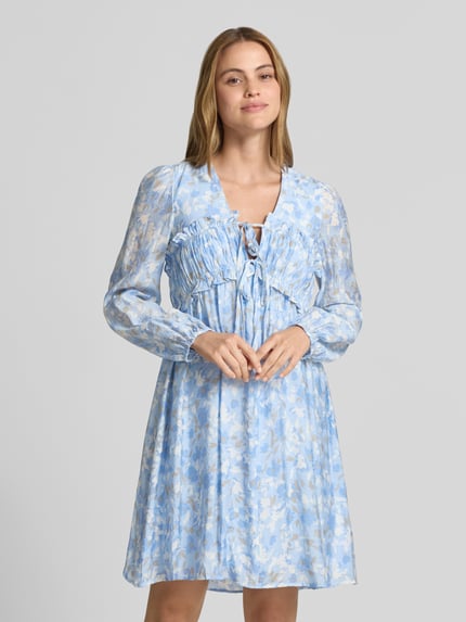 Frauenmodel trägt knielanges Kleid aus Viskose-Mix Modell 'Pipiana' von mbyM mit floraler Allover-Print, Rüschenbesatz, V-Ausschnitt, Langärmelig, elastische Ärmelabschlüsse, Taillierte Passform, zarte hellblaue und beigefarbene Töne, weich-fließende Textur, Ganzkörperansicht von vorne.