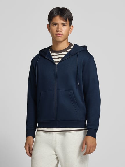 Męski model nosi Regular Fit Sweatjacke aus Baumwoll-Mix Model 'BRADLEY' von JACK & JONES, kolor granatowy, mieszanka bawełny, krój regular fit, kaptur z tunelem, oddzielna kieszeń kangurka, elastyczne wykończenia, długość tyłu w rozmiarze M: 67 cm, długość rękawów w rozmiarze M: 68 cm, zdjęcie całej sylwetki od przodu.