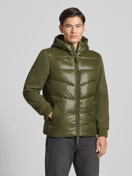 Männermodel trägt Pufferjacke mit Kapuze von CK CALVIN KLEIN in Olivgrün, unifarben, glänzend, Struktur, Regular Fit, Longline, Ganzkörperansicht von vorne.